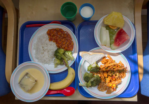 Folie 2 von 20: Two lunch trays at a primary school in London are served during a lunch break on Tuesday, May 6, 2014. The meal choice at right consists of pasta with fresh broccoli and slices of bread, and seasonal fresh fruit. At left are vegetable chili with rice and fresh broccoli, sponge cake with custard, and a banana. The drink options are milk and water. (AP Photo/Sang Tan)