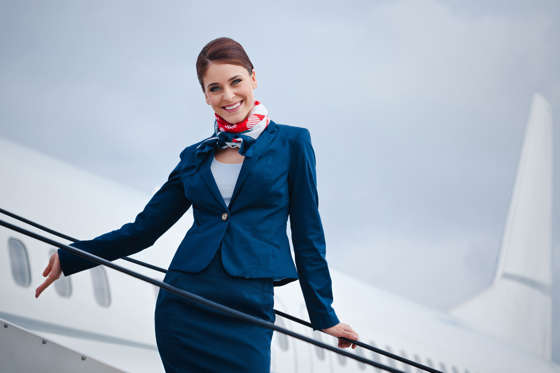 Slide 1 of 8: Outdoor portrait of a beautiful flight attendant standing on the aircraft stairs and smiling at the camera.