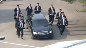 a group of people standing on top of a car: Jogging security guards flank Kim Jong Un's limo
