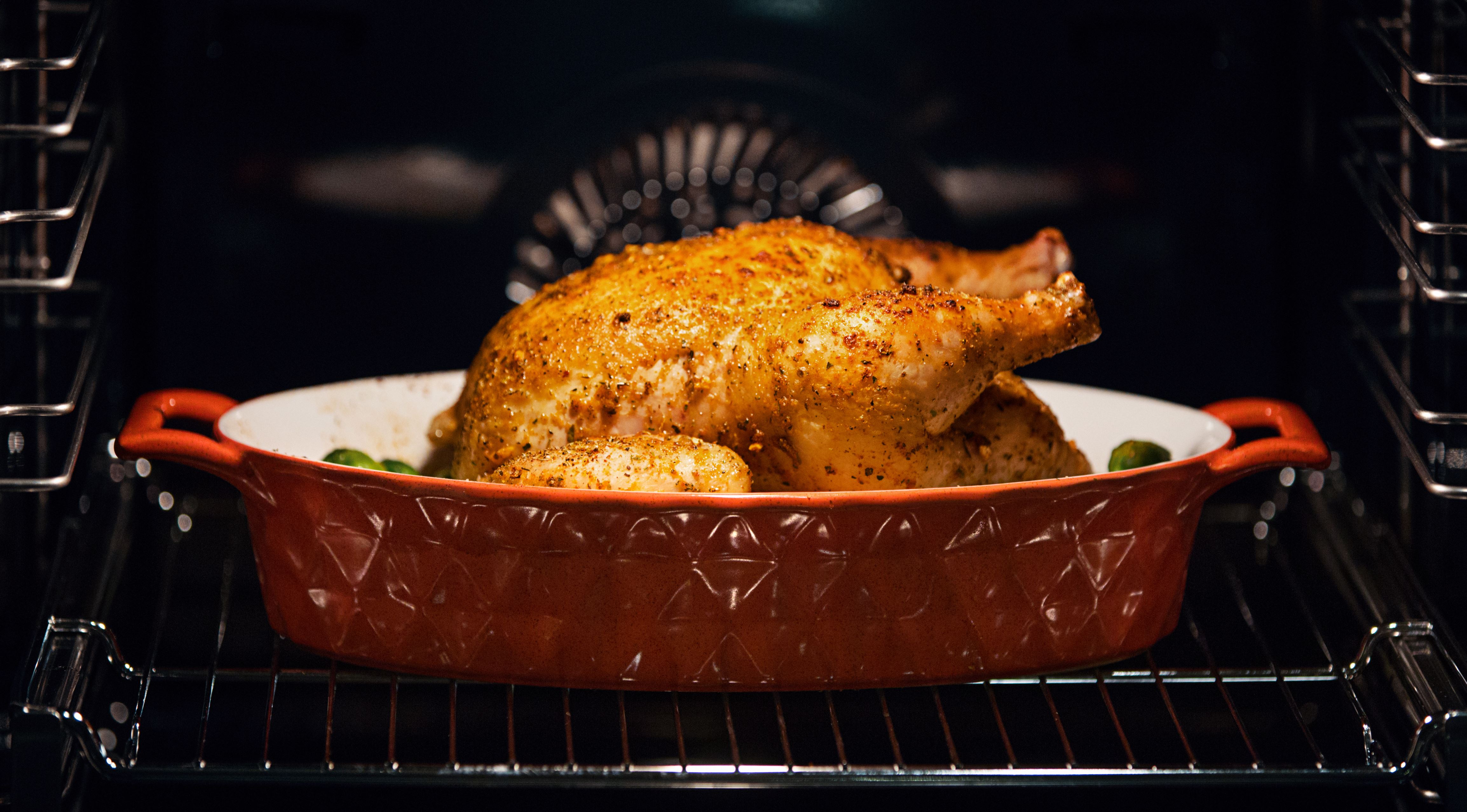 Closeup side view of a chicken in a clay pot roasting in an oven. It's almost  done with golden crispy skin and some green vegetables.