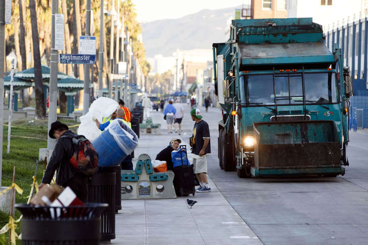 people sit as sanitation workers empty trash cans during morning cleanup of homeless encampments on the Venice Beach boardwalk in Los Angeles, California, U.S., on Friday, Oct. 9, 2015.