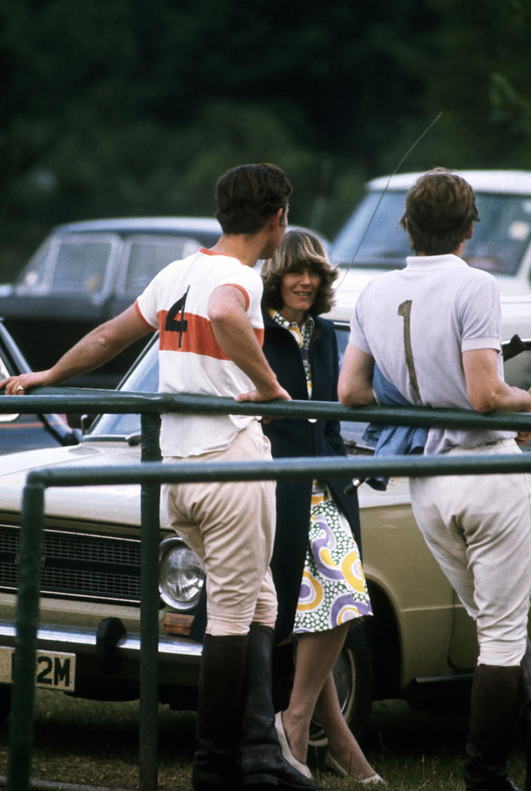 En 1972, en medio de un partido de polo, **Carlos III** del Reino Unido y la actual **reina consorte** comparten de una charla. Camilla optó por un vestido midi de estampado vibrante, gabardina negra y stilettos. 
