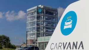 Carvana (CVNA stock) logo on white object in foreground as well as a high-rise building in the background