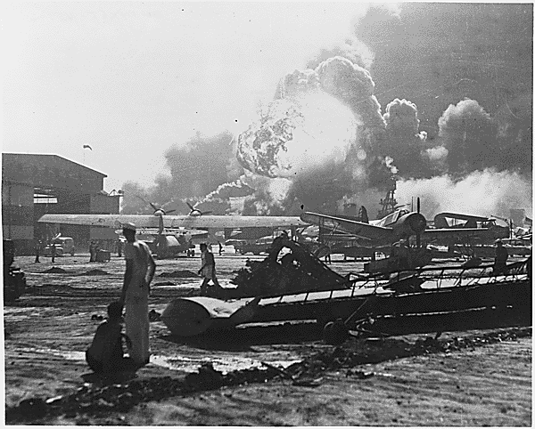 <p>US Navy personnel standing among the aircraft and equipment damaged during the attack. Smoke and flames can be seen in the distance, rising into the air from the battleships that were targeted by Japanese aircraft.</p>