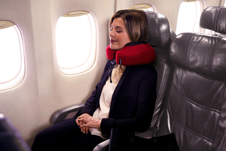 Woman using red Cabeau Evolution S3 Travel Pillow in window seat of plane.