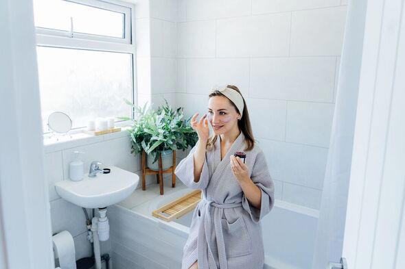Young woman in bathrobe looking in the mirror and applying facial natural cosmetic clay mask on her face in bathroom. Cosmetic procedures for skin car