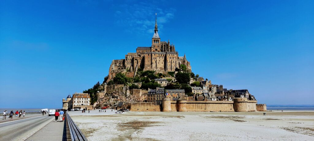 <p><em>By Faith of XYUandBEYOND</em></p> <p>Mont Saint Michel is located on a small island just off the coast of Normandy and is a stunning UNESCO World Heritage site. <a href="https://xyuandbeyond.com/visiting-mont-st-michel/" rel="noreferrer noopener">Mont Saint Michel</a> is a medieval Abbey and village constructed over 1300 years ago.</p> <p>To get to the island you take a free shuttle or you can walk or bike it. Technically it is free to visit the Mont, but parking will cost you €14.90 and there are charges to visit the small museums on the Mont itself along with a €10 fee to enter the Abbey.</p> <p>Mont St. Michel contains a small village in which live around 50 people and a medieval monastery. It is only 17 acres and sits around a kilometre from the shore.</p> <p>The Village of Mont Saint Michel starts right at the entrance and from there it's all uphill. At the top of the main street, starts the “Grand Degre” 350 steps which lead up to the Abbey.</p> <p>When you see the Mont close up you will spot ramparts that circle the island and a 3 tiered assembly of buildings from the 13th century known as La Merveille (The Wonder) that rise up to the abbey's pointed spire. On the second terrace of La Merveille is Mont-St-Michel's largest and most beautiful space, a 13th-century hall known as the Salle des Chevaliers. Crowning the mount's summit is the spellbinding Eglise Abbatiale church. Only 350 or so steps to reach the Abbey and when you get there the entry ticket will cost €10 euros.</p>