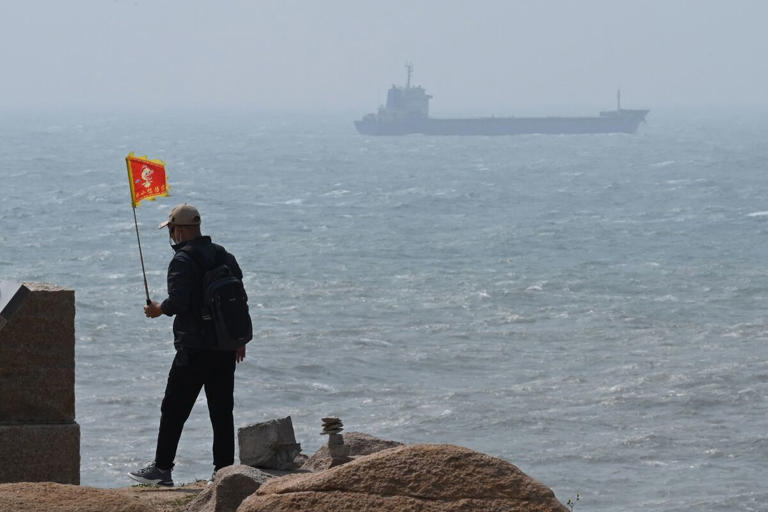 Un hombre camina con una bandera, mientras un barco pasa detrás de él en la isla de Pingtan, el punto más cercano a Taiwán, en la provincia de Fujian. Foto: AFP