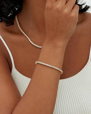 Person wearing a white ribbed tank top showcases a sparkling gold necklace and bracelet, both with diamond embellishments