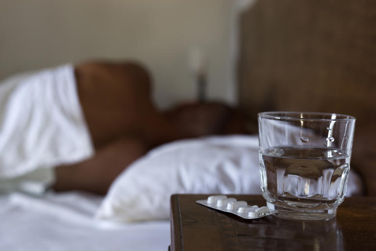 Water glass, a pack of tablets / pills and a man sleeping in a bed in the blurred background