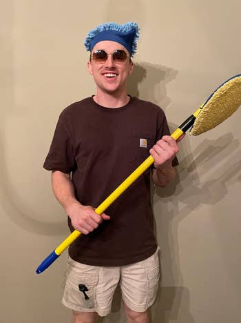 Person smiling and posing with sunglasses and casual clothes, holding a cleaning mop, possibly for a fun home cleaning or DIY article