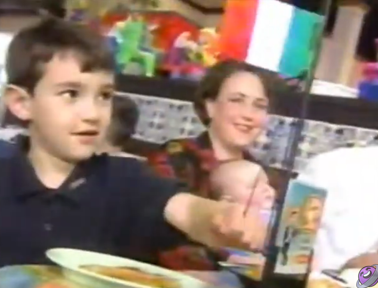 Boy holding Mexican flag at a table with family, including a woman holding a baby. Toy decor is visible in the background