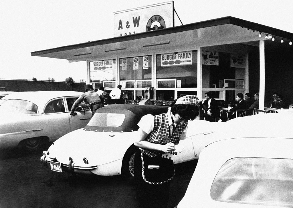 People gather outside an A&W restaurant in a vintage setting, with classic cars parked nearby