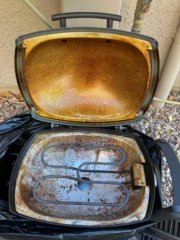 Outdoor electric grill with lid open, showing a greasy interior and rusted heating element. The grill is set on a rocky ground