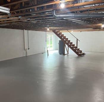 Unfinished basement with exposed beams, concrete floor, water heater, and wooden stairs leading to an exit door with natural light