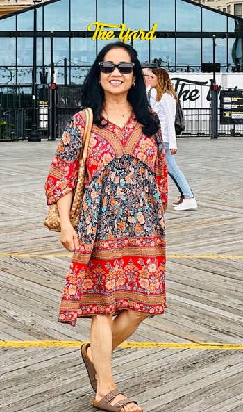 Person in a patterned bohemian-style dress standing outdoors in front of a building with a sign saying