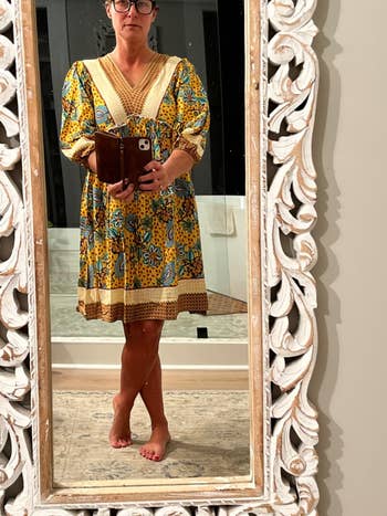 Woman stands barefoot in front of a mirror, wearing a floral-patterned dress with puffed sleeves and intricate trim, holding a phone case