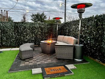 Outdoor patio set with gray wicker seats around a fire pit on a tiled surface, flanked by tall patio heaters, surrounded by tall hedges