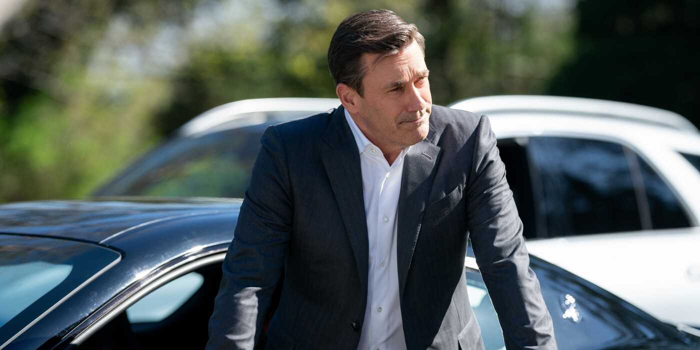 Coop, wearing black jacket and white shirt, leaning in front of a car in Your Friends & Neighbors