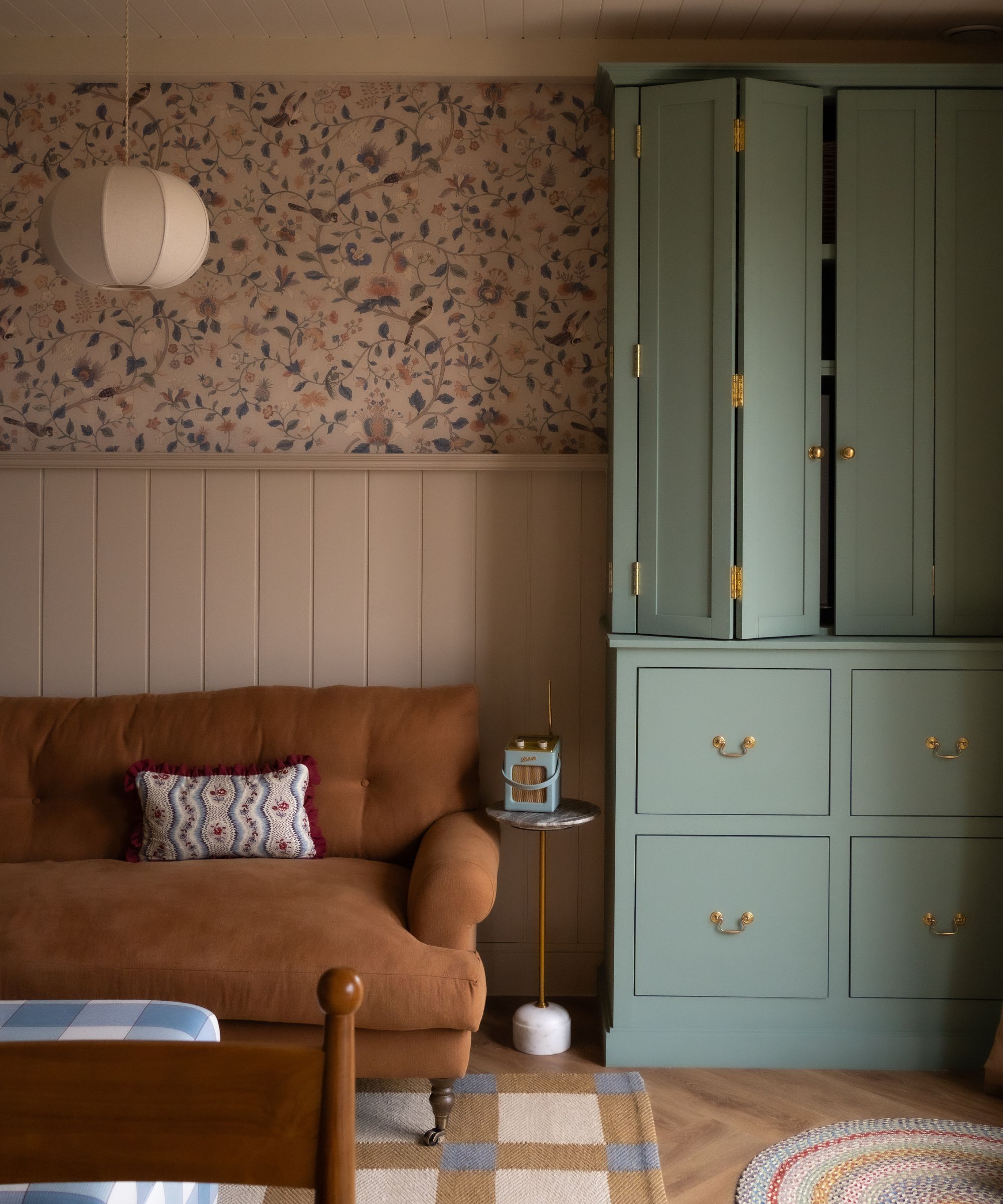small snug area with paneled walls and half floral wallpaper, a tan sofa and a blue custom cabinet that folds out to reveal the TV