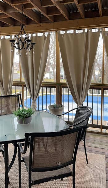 Covered patio with a glass table, chairs, and curtains, overlooking a pool in a backyard setting