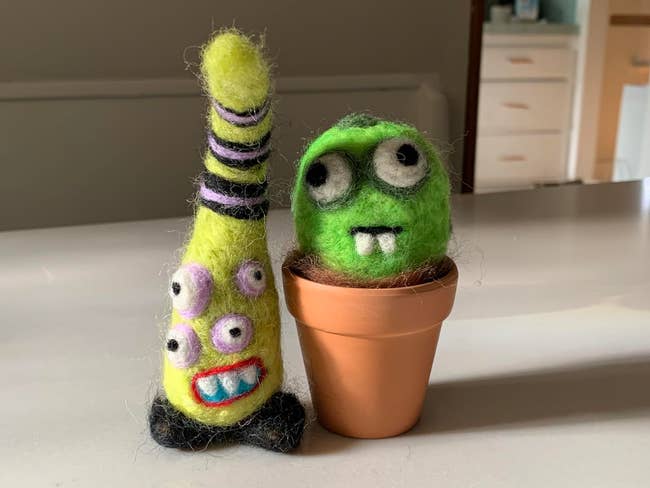 Two whimsical felt figures: one tall, cone-shaped with stripes and eyes, and one round, green with big eyes in a small clay pot. Quirky decor pieces