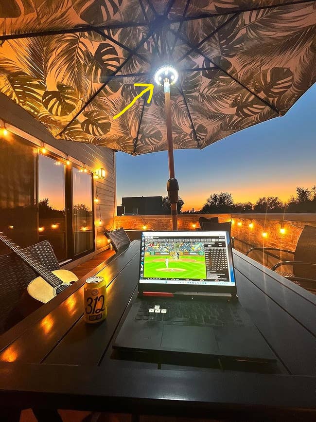 Outdoor patio setup with a laptop showing a game, a guitar, and a drink on the table under a decorative umbrella at sunset