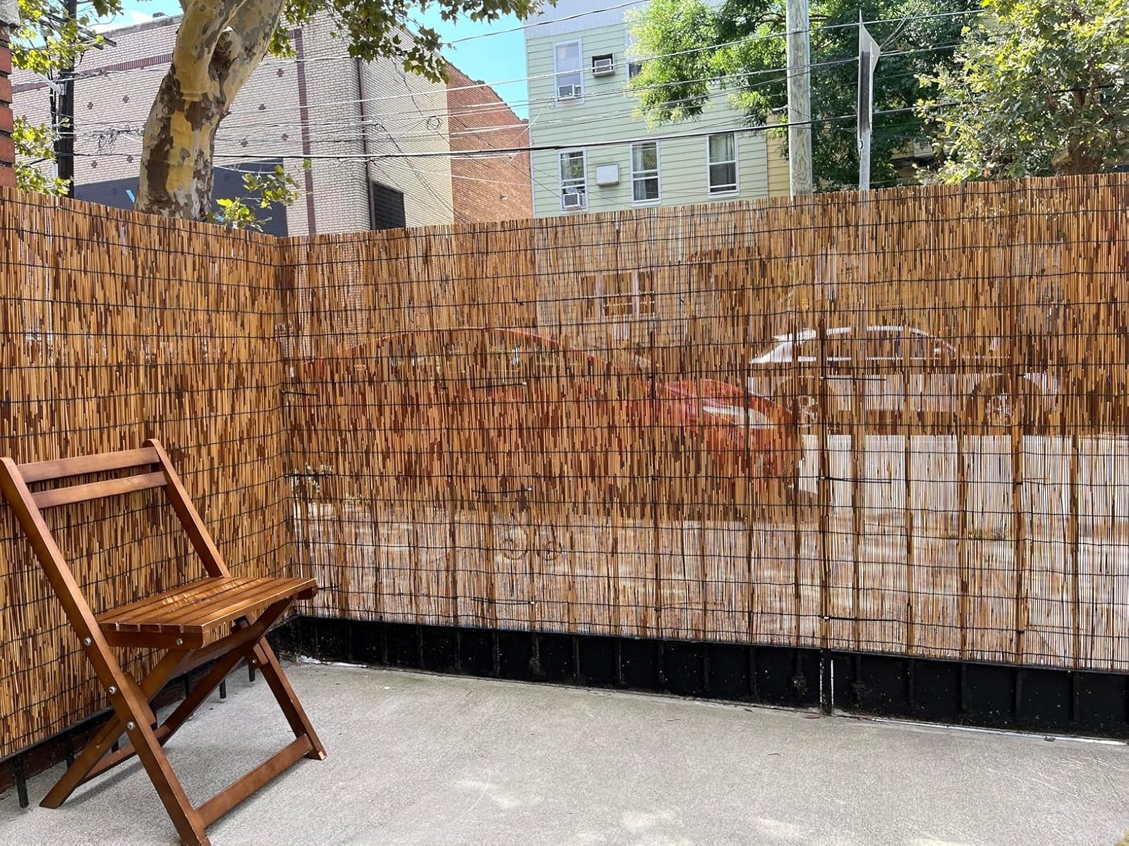 Wooden chair on a patio next to a tall bamboo fence, with buildings and parked cars visible through gaps in the fence