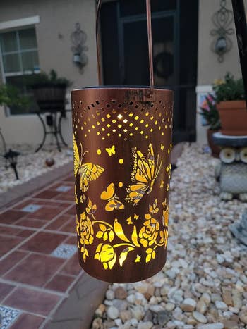 Decorative lantern with butterfly and floral cutouts, casting a warm glow, hangs on a patio surrounded by rocks and a tiled path