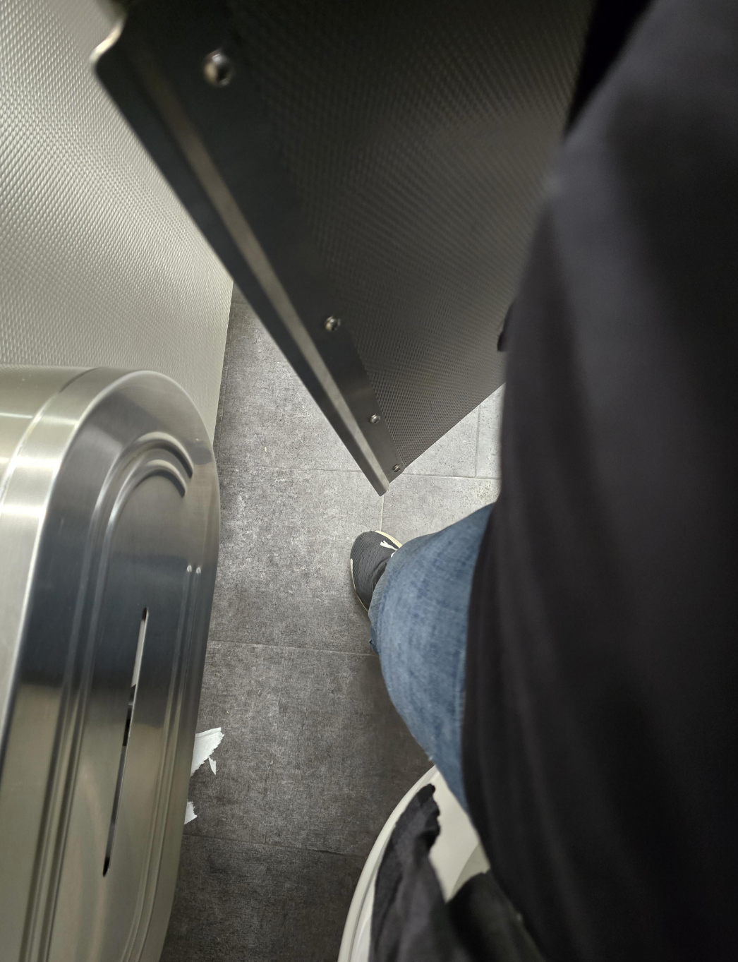 Person inside a restroom stall, partially visible, with one leg in jeans and sneakers. Door slightly ajar, showing part of a metal bin