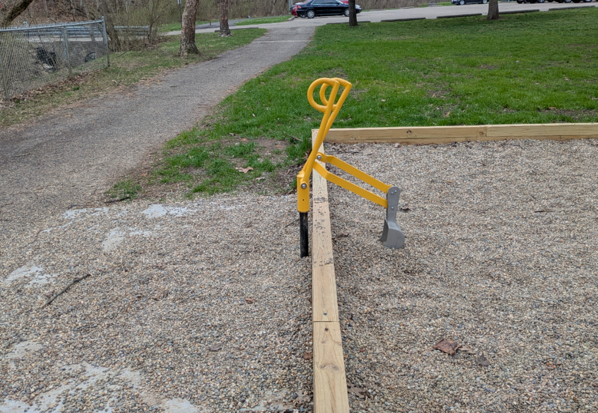 Seesaw in a playground with one end stuck in gravel, rendering it unusable. Grass and a path are in the background