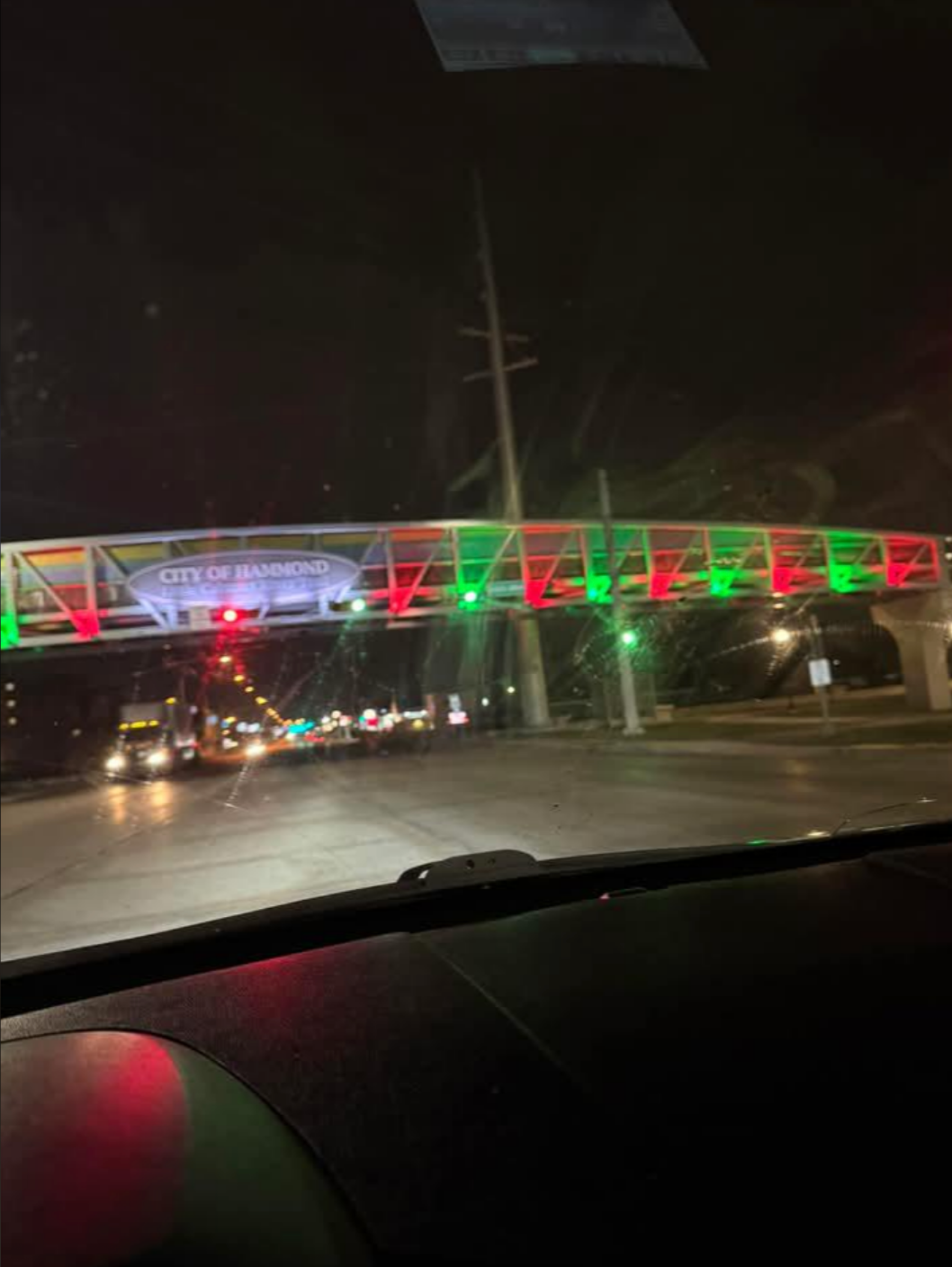Night view of an illuminated pedestrian bridge over a road, with "City of Hammond" sign and traffic lights visible
