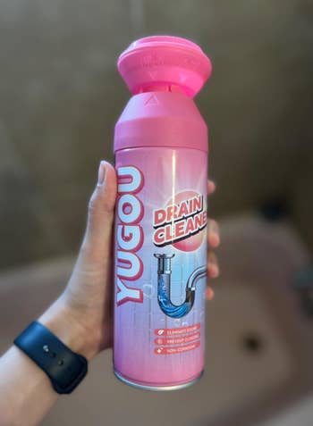 Person holding a Yugou Drain Cleaner bottle, featuring a pipe graphic and label highlighting odor elimination and clog prevention