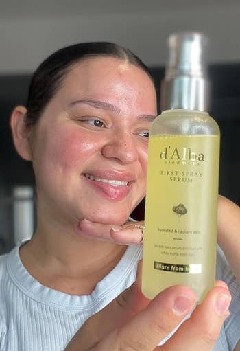 Person holding a bottle of d'Alba spray serum, smiling and showcasing the product for skincare use