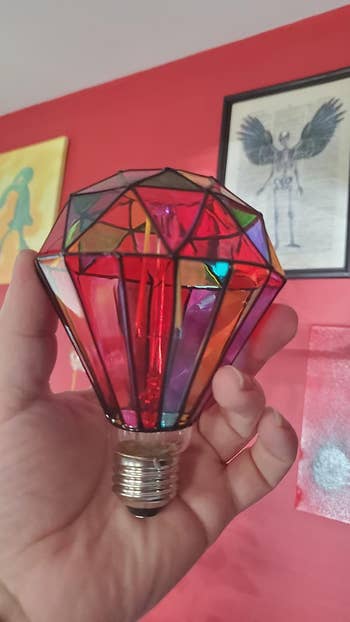 Stained glass-style light bulb held in a hand, surrounded by eclectic artwork on a red wall