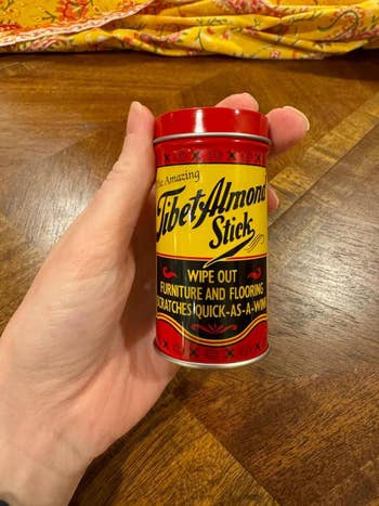 Hand holding a Tibet Almond Stick, a scratch remover for furniture and flooring. The cylindrical packaging features vintage-style text and design
