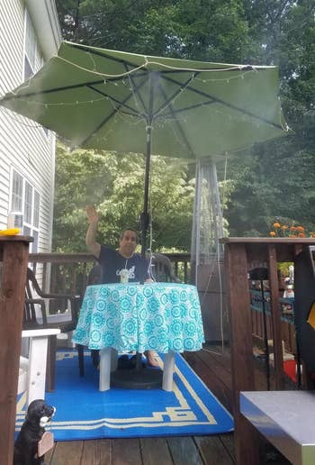a reviewer sits on their patio under an umbrella with the orbit outdoor mist cooling system attached to it and spraying mist