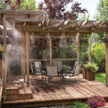Outdoor patio with a wooden pergola, dining set, and misting system, surrounded by lush greenery and flowers, creating a refreshing retreat