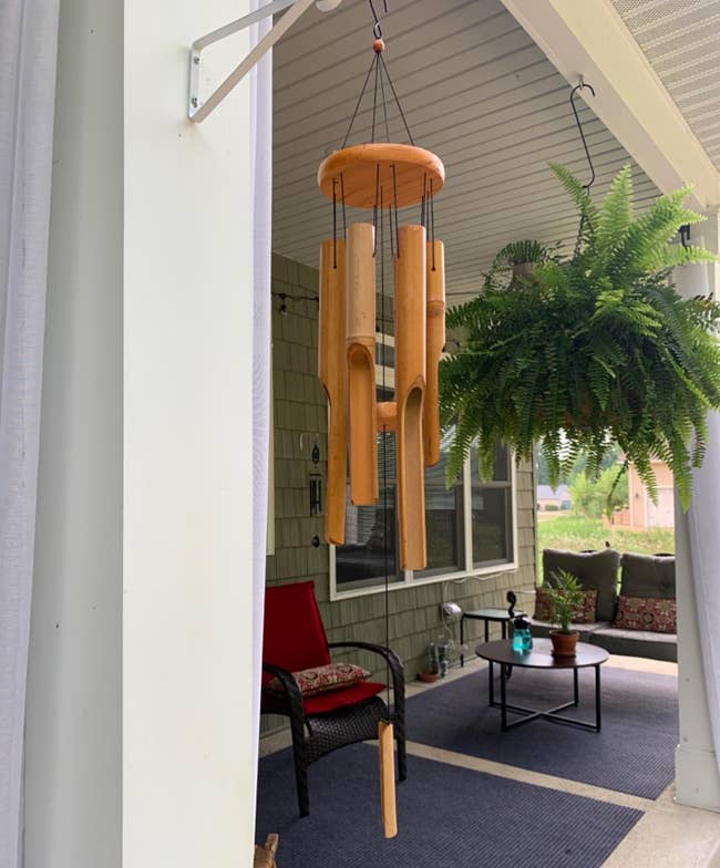 A porch with a hanging wooden wind chime and a large fern. There is outdoor seating with cushions and a small table with a bottle on it