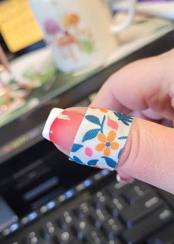 A hand with a floral-patterned bandage on the thumb over a French manicure, with blurred office items in the background