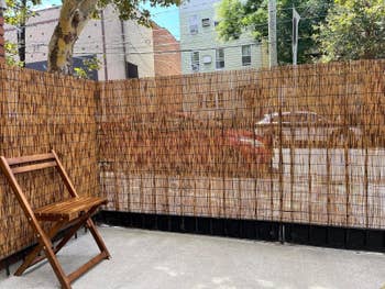 Wooden chair on a patio next to a tall bamboo fence, with buildings and parked cars visible through gaps in the fence