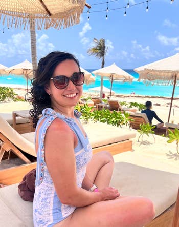 Person relaxing on a beach lounge chair under a straw umbrella, wearing sunglasses and a light-patterned outfit, with the ocean in the background