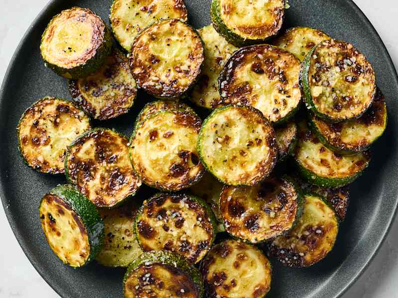 Sautéed zucchini rounds with a golden-brown crust, garnished with seasoning on a dark plate.
