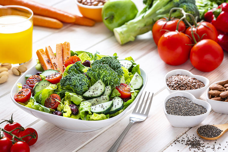 Vegan food: healthy salad plate shot on white table. The salad plate is surrounded by fresh organic multicolored fruits, vegetables, seeds and nuts like broccoli, tomatoes, carrots, lettuce, green apple, cucumber, pinto beans, pistachio, almonds, chia seeds, brown flax seeds among others. High resolution 42Mp studio digital capture taken with SONY A7rII and Zeiss Batis 40mm F2.0 CF lens