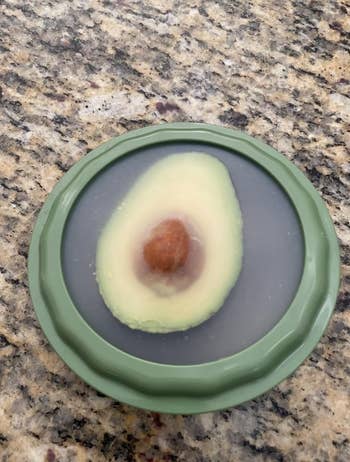 Half an avocado with a pit is stored cut-side up in a green silicone food saver on a granite countertop