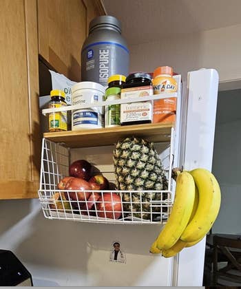 Kitchen shelf with supplements, a large water bottle on top, and a basket holding apples, a pineapple, and bananas hanging outside