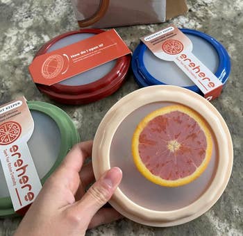 Four silicone food covers in various colors are shown, each featuring a realistic slice of fruit at the center. A hand holds one cover