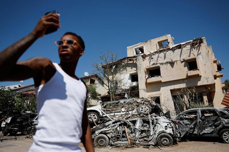 A man uses his smartphone in front of destroyed cars at an impact site following missile attack from Iran on Israel, in Rishon LeZion, Israel, June 14, 2025.