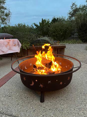 A firepit with star and moon cutouts burns brightly on a patio, surrounded by outdoor seating and foliage