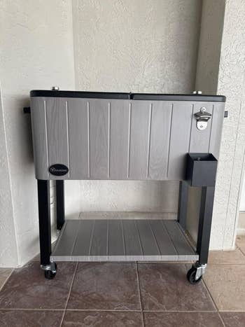 A large wheeled cooler on a stand with a bottle opener and storage shelf below, placed against a wall